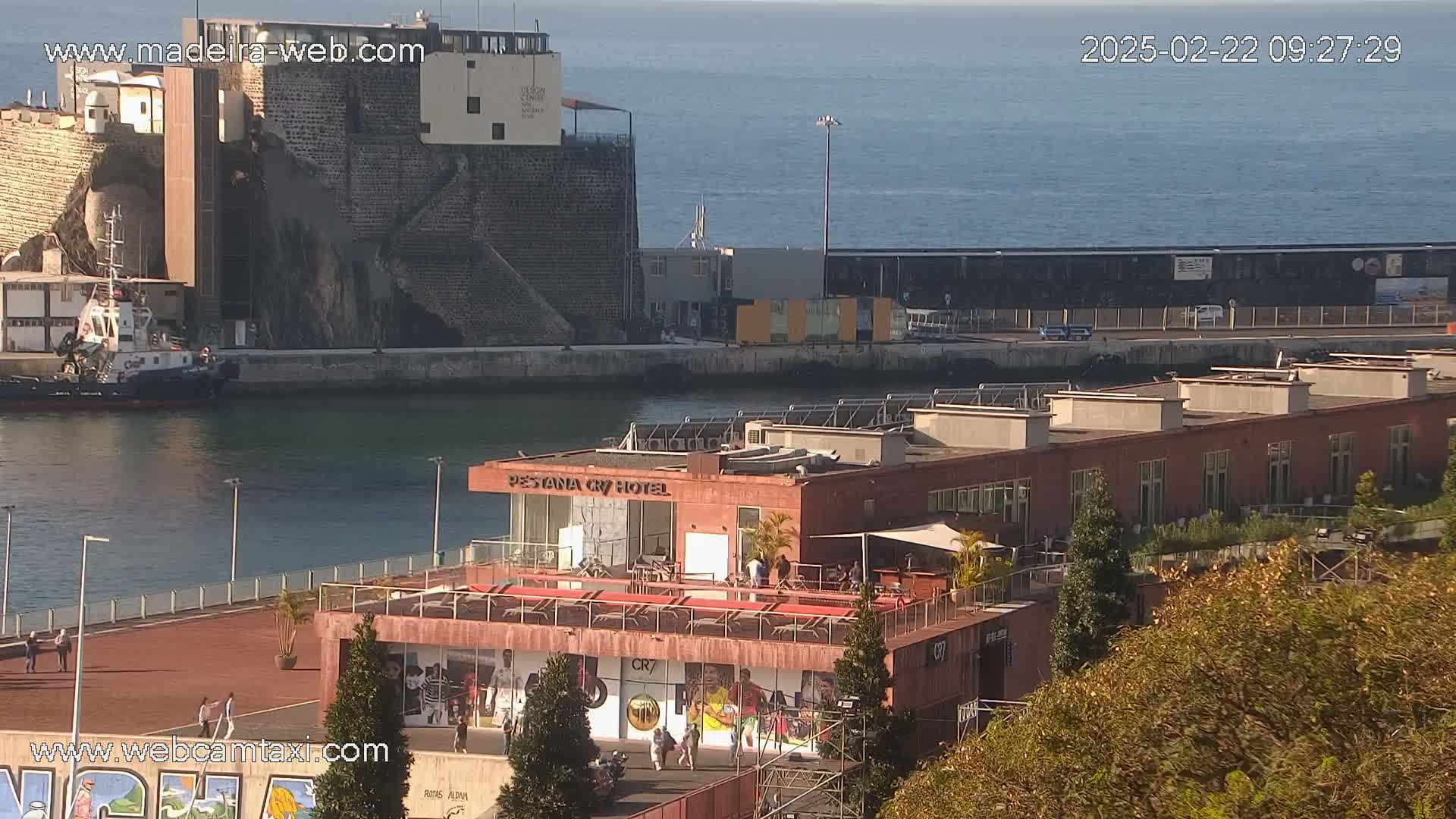 A high-angle view of a harbor on a sunny day, showing a tugboat, a brick hotel building with a rooftop patio, and other buildings along the waterfront.