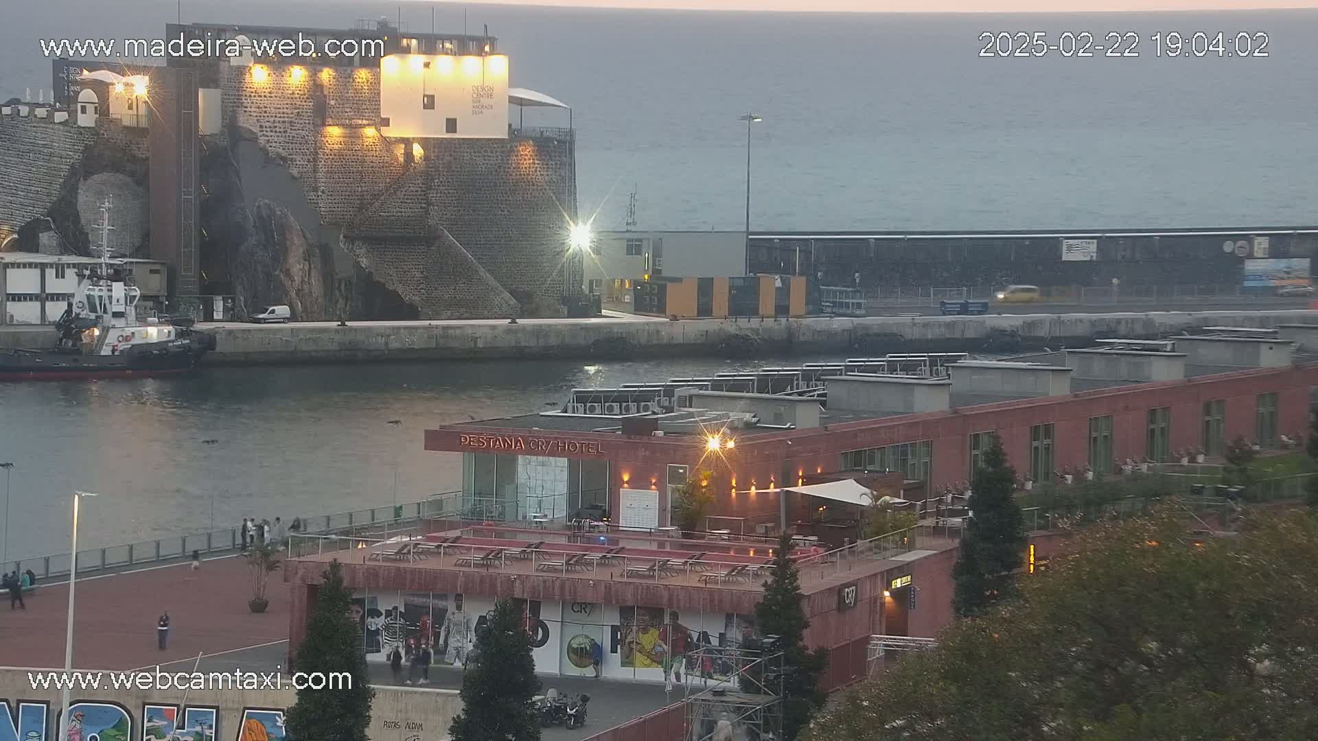 A harbor scene at dusk shows a tugboat in the water, a stone structure with lights, a low-rise building with outdoor seating, and a calm body of water.