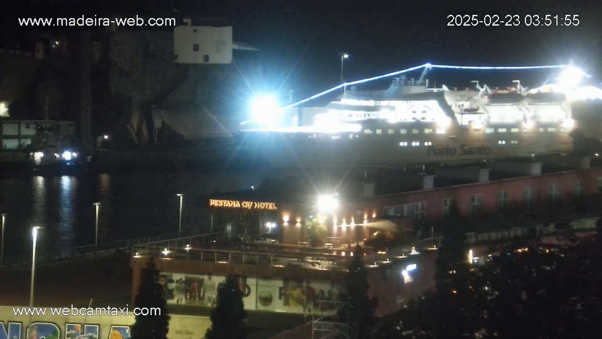 A large, brightly lit ferry is docked at night near a hotel and other buildings by a body of dark water.