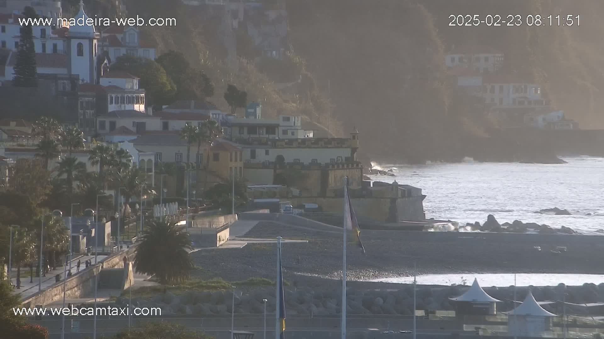 A coastal town with buildings perched on a hillside overlooking a pebble beach and a somewhat choppy ocean under hazy sunlight.