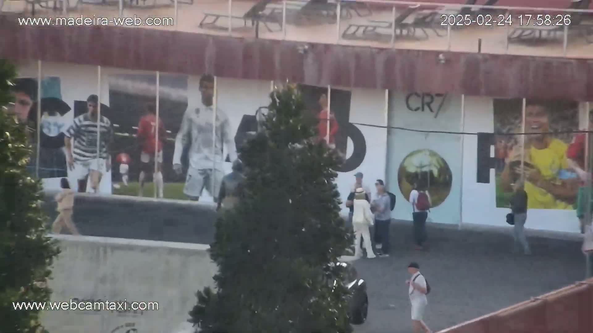 A wall with large soccer player images is visible outdoors on a partly sunny day, with several people walking near it.