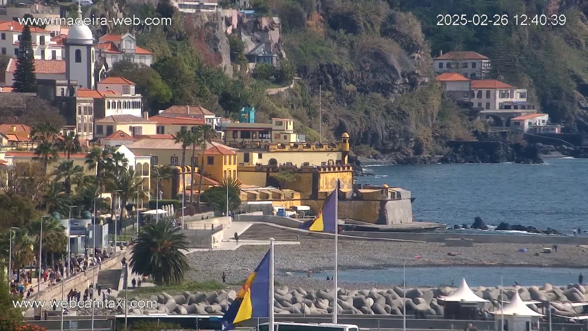A coastal town with colorful buildings and a stone fortress sits on a rocky shoreline next to a calm ocean under a sunny sky.