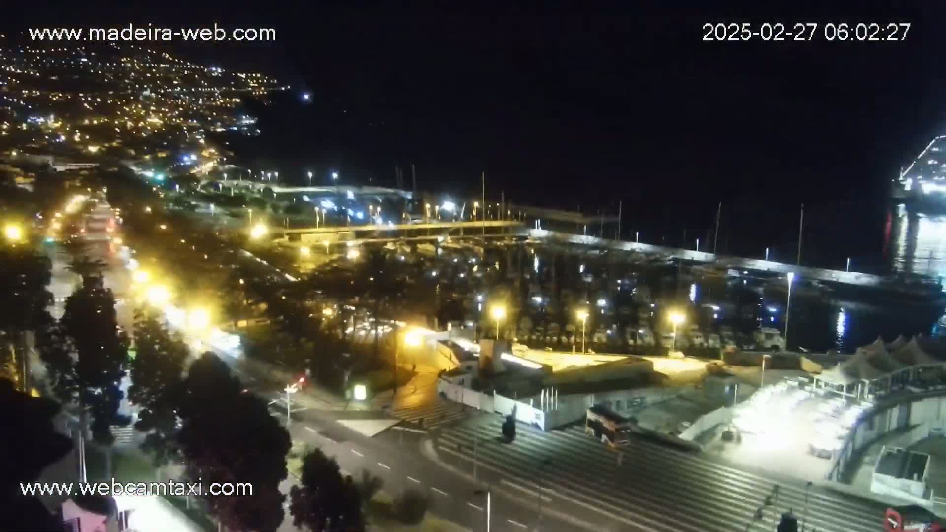 A nighttime, high-angle view shows a marina filled with boats, a roadway with traffic, and a city illuminated with lights in the background.