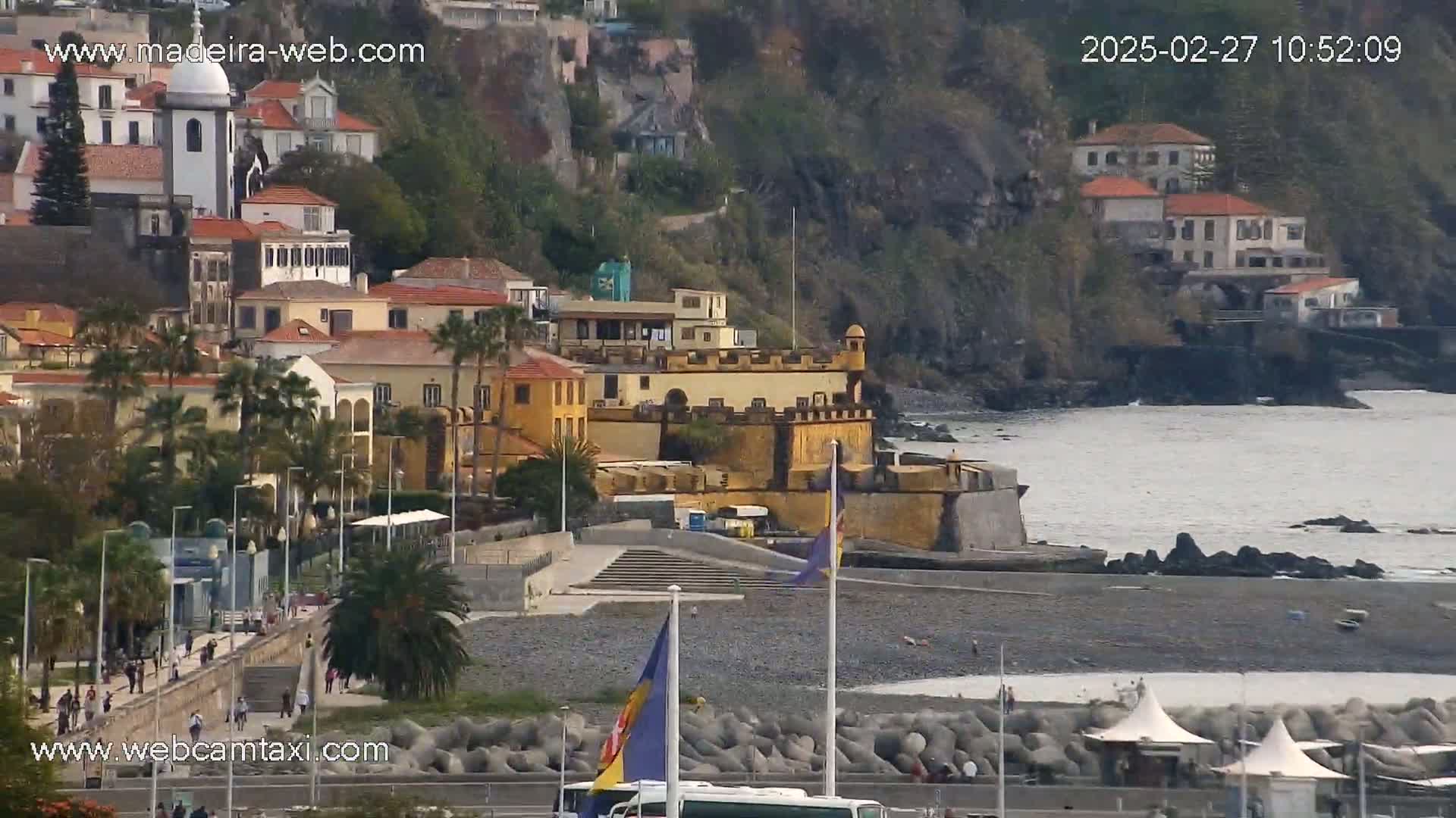 A coastal town with a yellow fort sits on a rocky shoreline, with calm water visible in the distance on an overcast day.