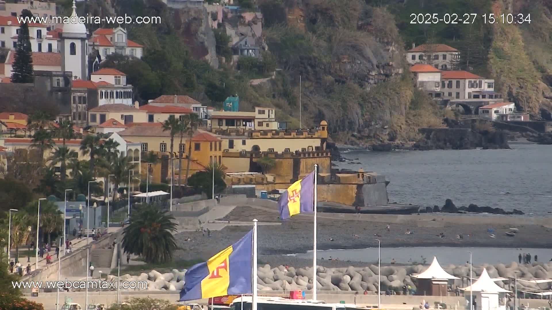A coastal town with colorful buildings and a fortress overlooks a dark-sand beach on a sunny day.