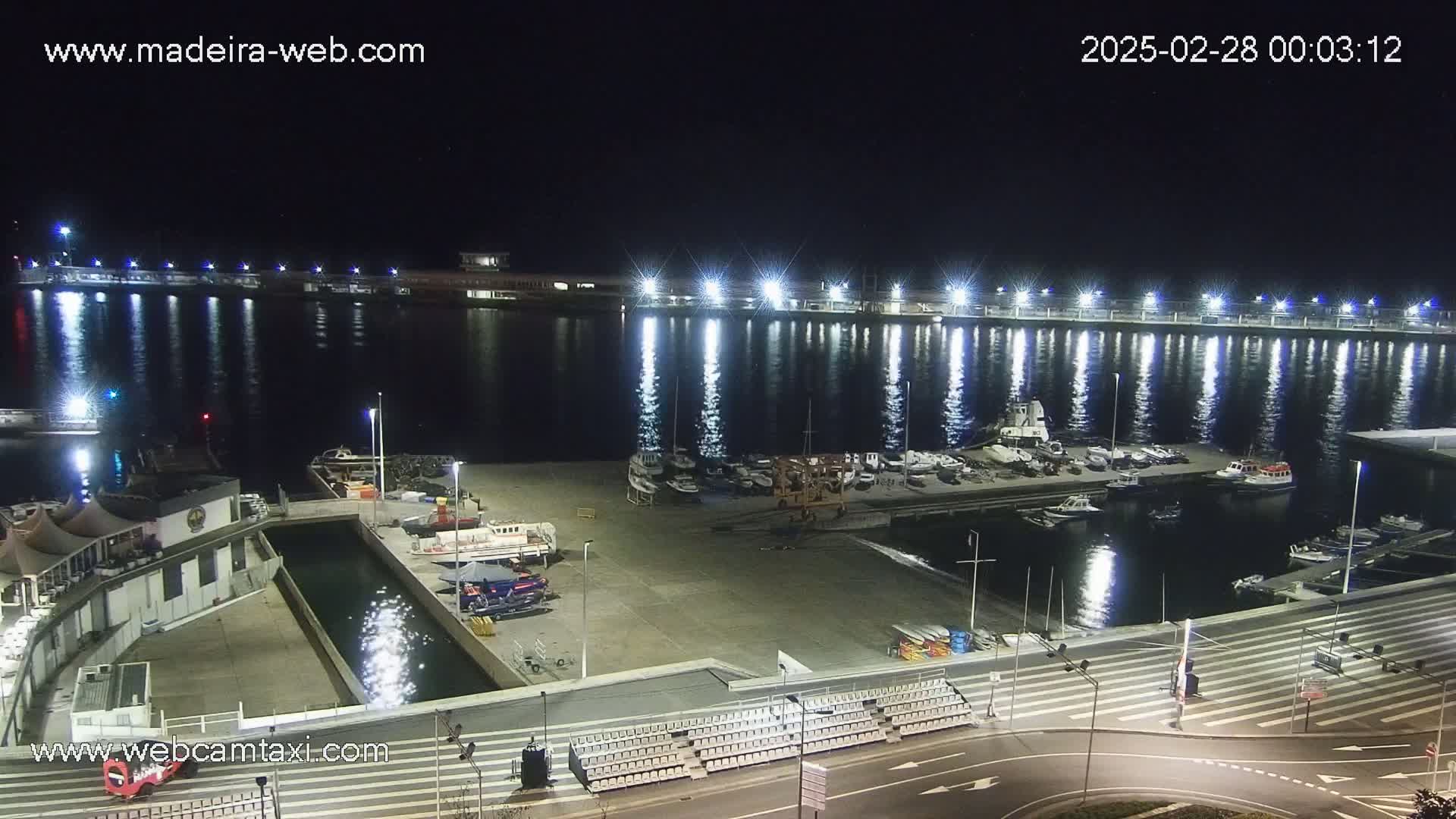 A nighttime view of a marina filled with boats, illuminated by bright lights reflecting on calm water.