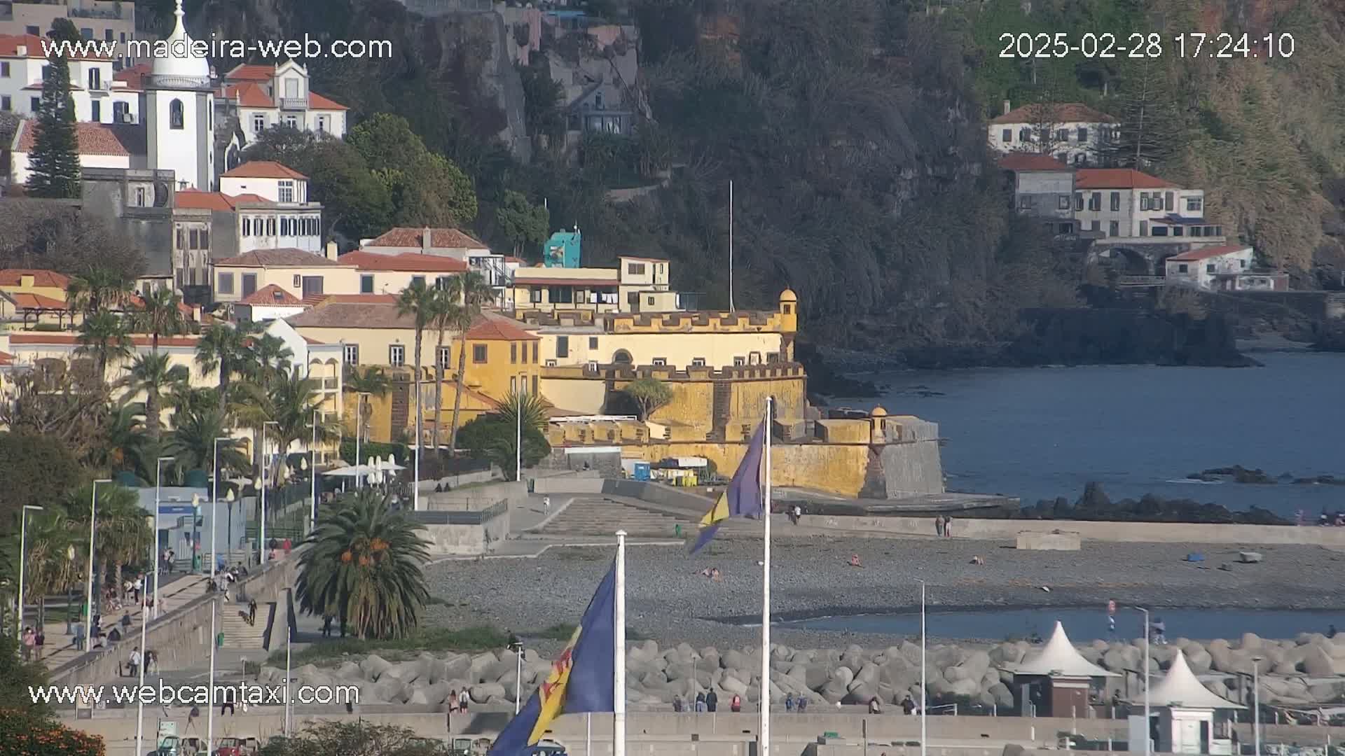 A coastal town with yellow buildings, a fortress, and a pebble beach is seen on a sunny day.