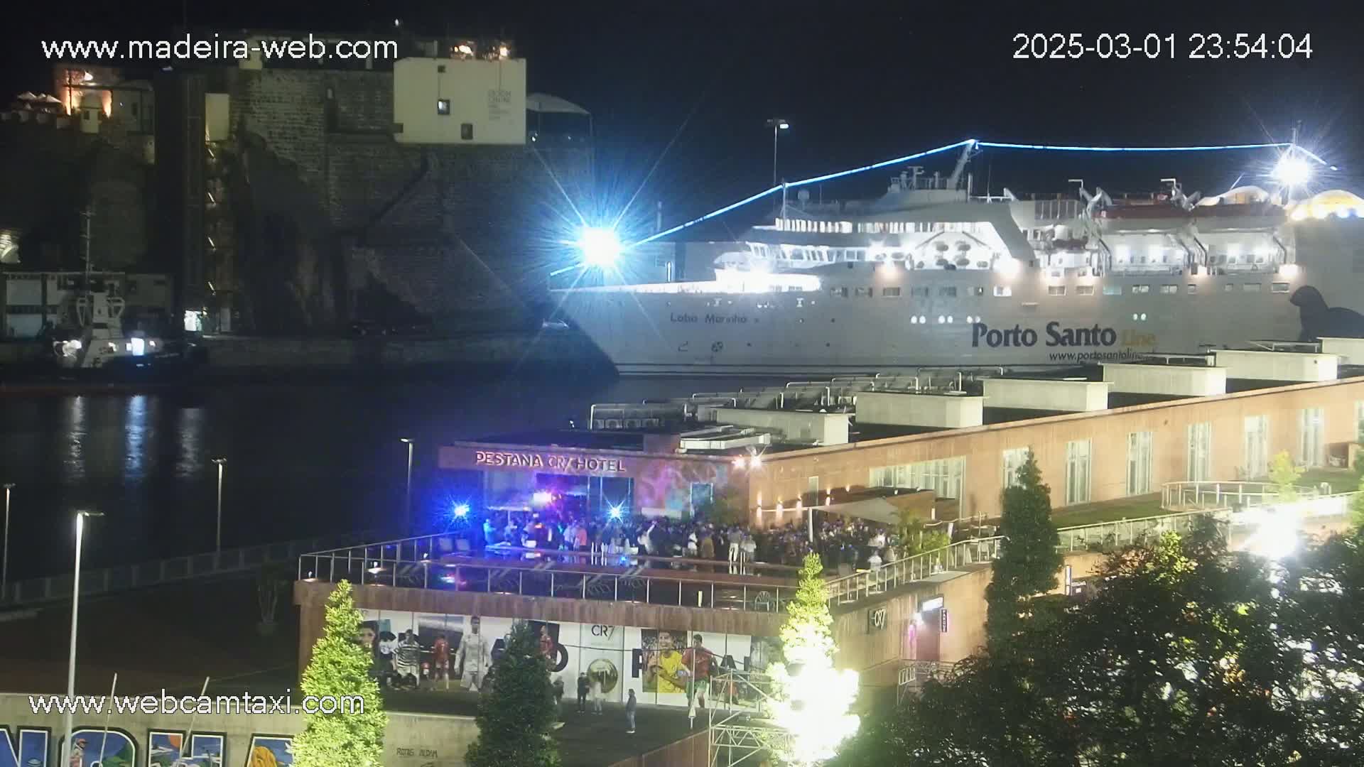 A large ferry is docked at a harbor at night, with a crowd of people gathered on a rooftop patio of a building nearby.