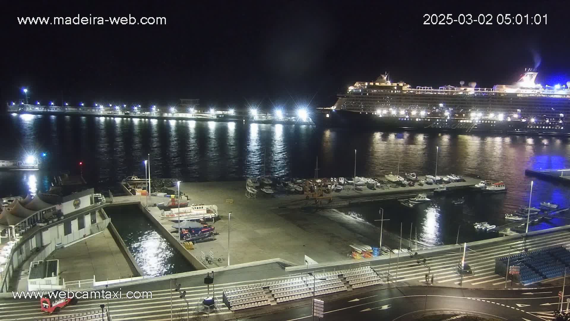 A large cruise ship is docked at a harbor at night, with smaller boats moored nearby and empty bleachers on the waterfront.