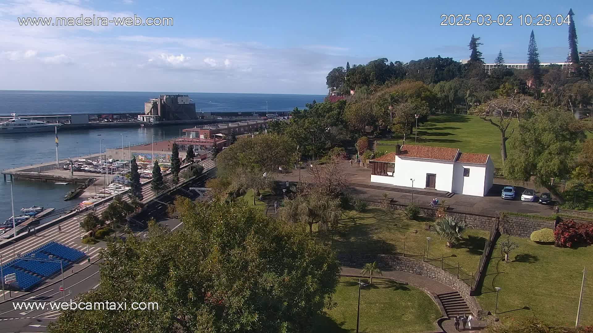 An elevated view of a harbor, marina, and park with a small white building, under sunny skies.