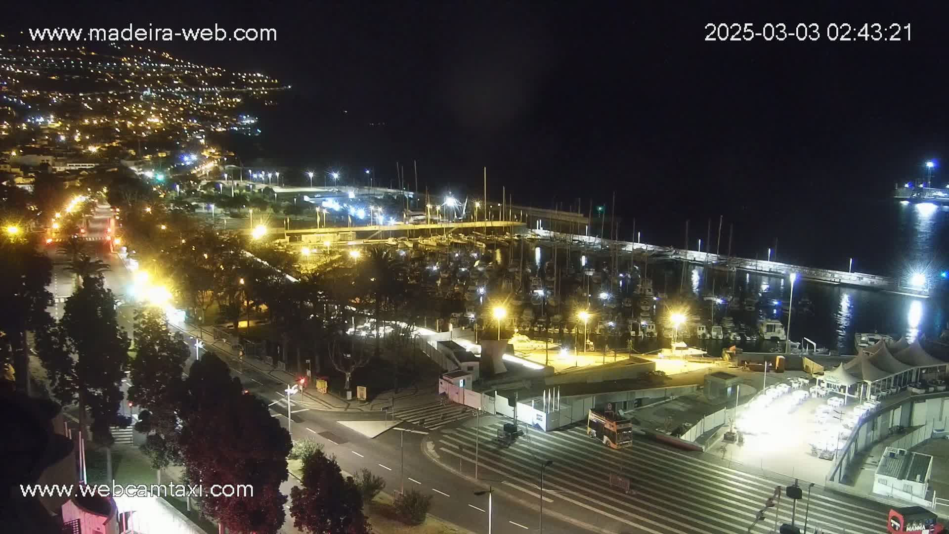 A nighttime aerial view shows a marina filled with boats, a roadway with traffic, and a hillside town illuminated with lights.