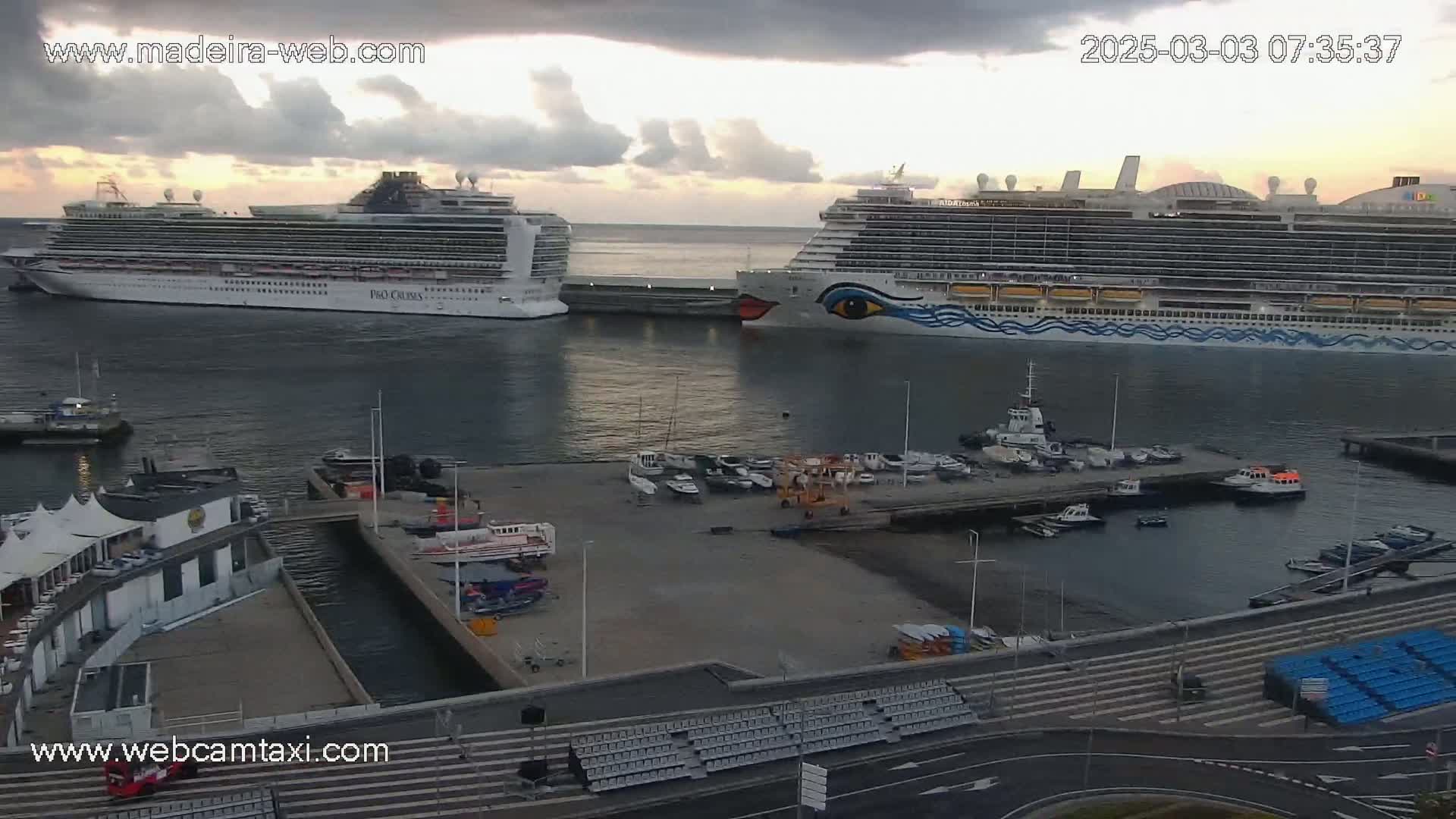 Two large cruise ships are docked in a harbor under a partly cloudy sky.
