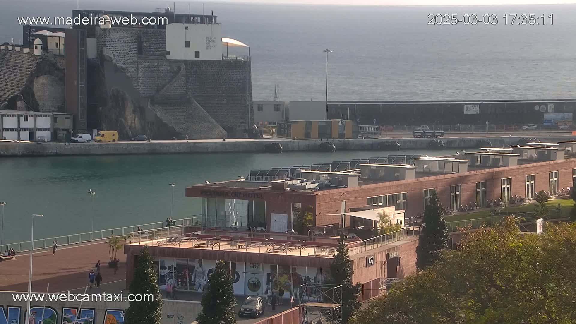 A harbor scene on a partly cloudy day shows a modern building with a rooftop patio overlooking a calm body of water, beyond which are older stone structures and a distant ocean.