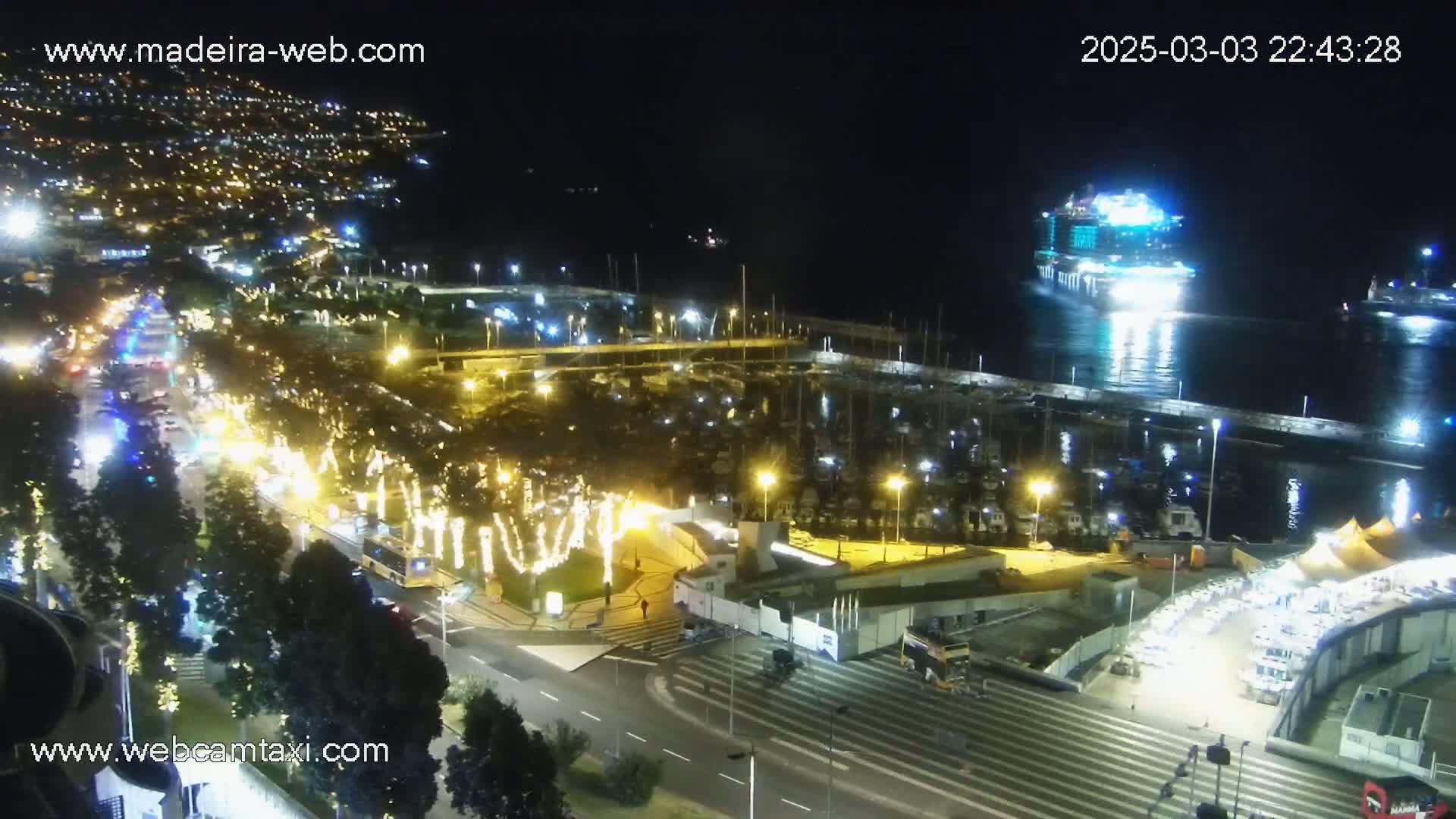 A brightly lit marina and city at night, with a large cruise ship in the harbor, under a clear, dark sky.