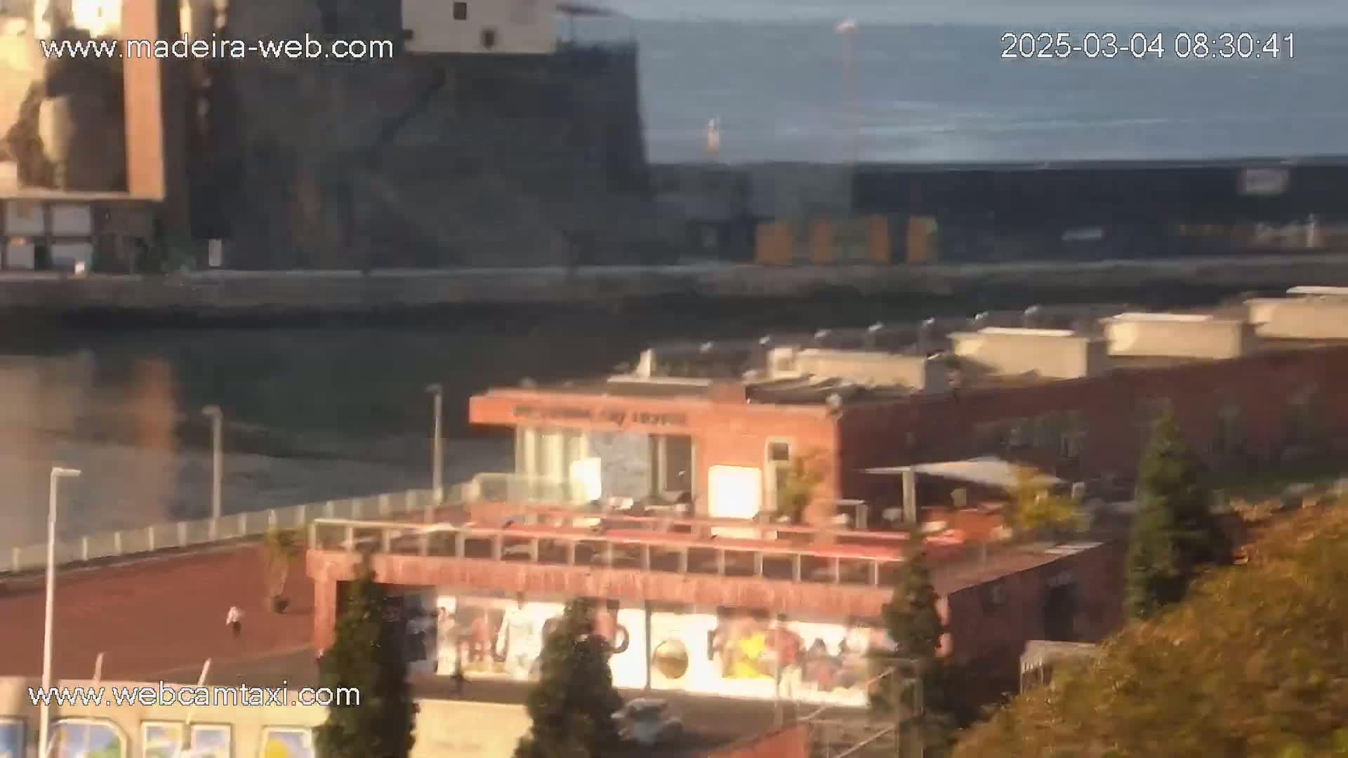 A high-angle, long shot shows a waterfront scene on a calm, sunny day, featuring a building with a rooftop patio overlooking a waterway and other buildings beyond it.