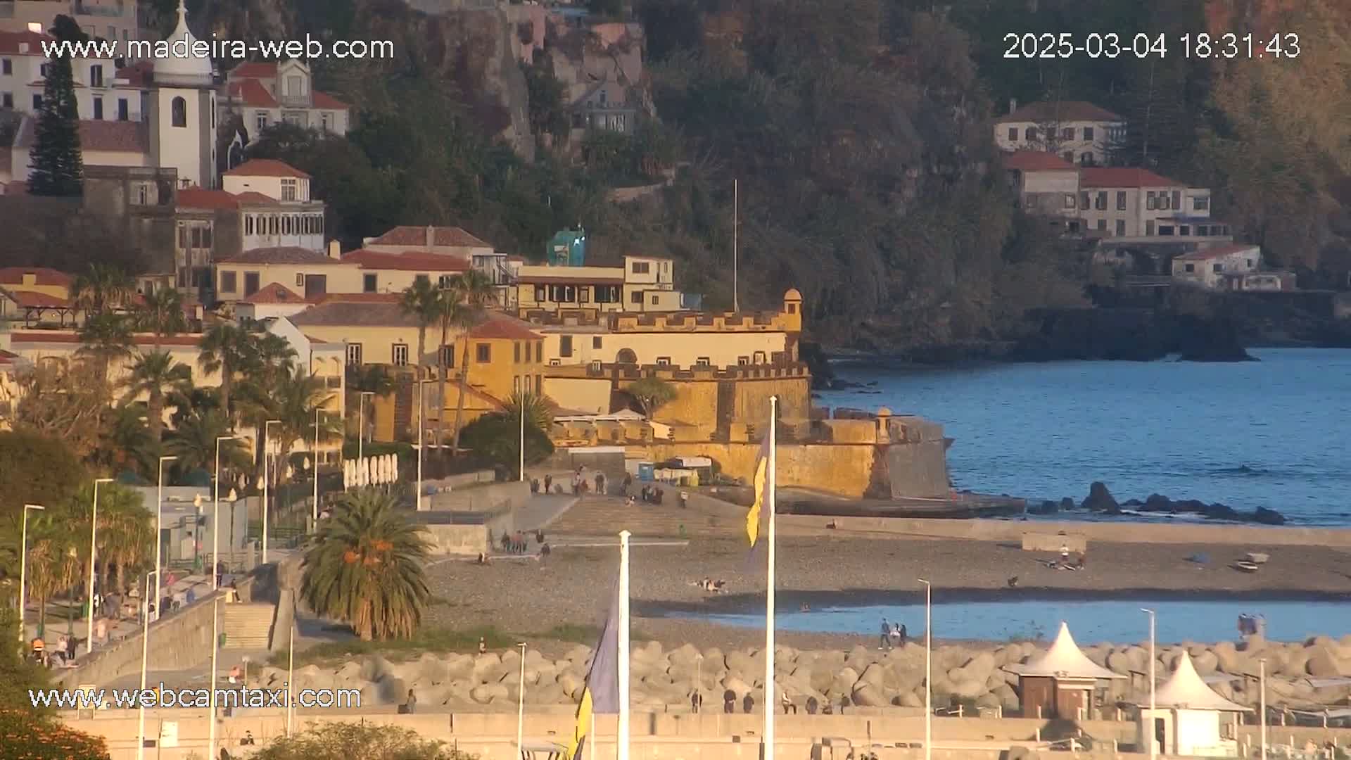 A coastal town with a stone fortress overlooks a pebble beach and calm ocean under a late afternoon sun.