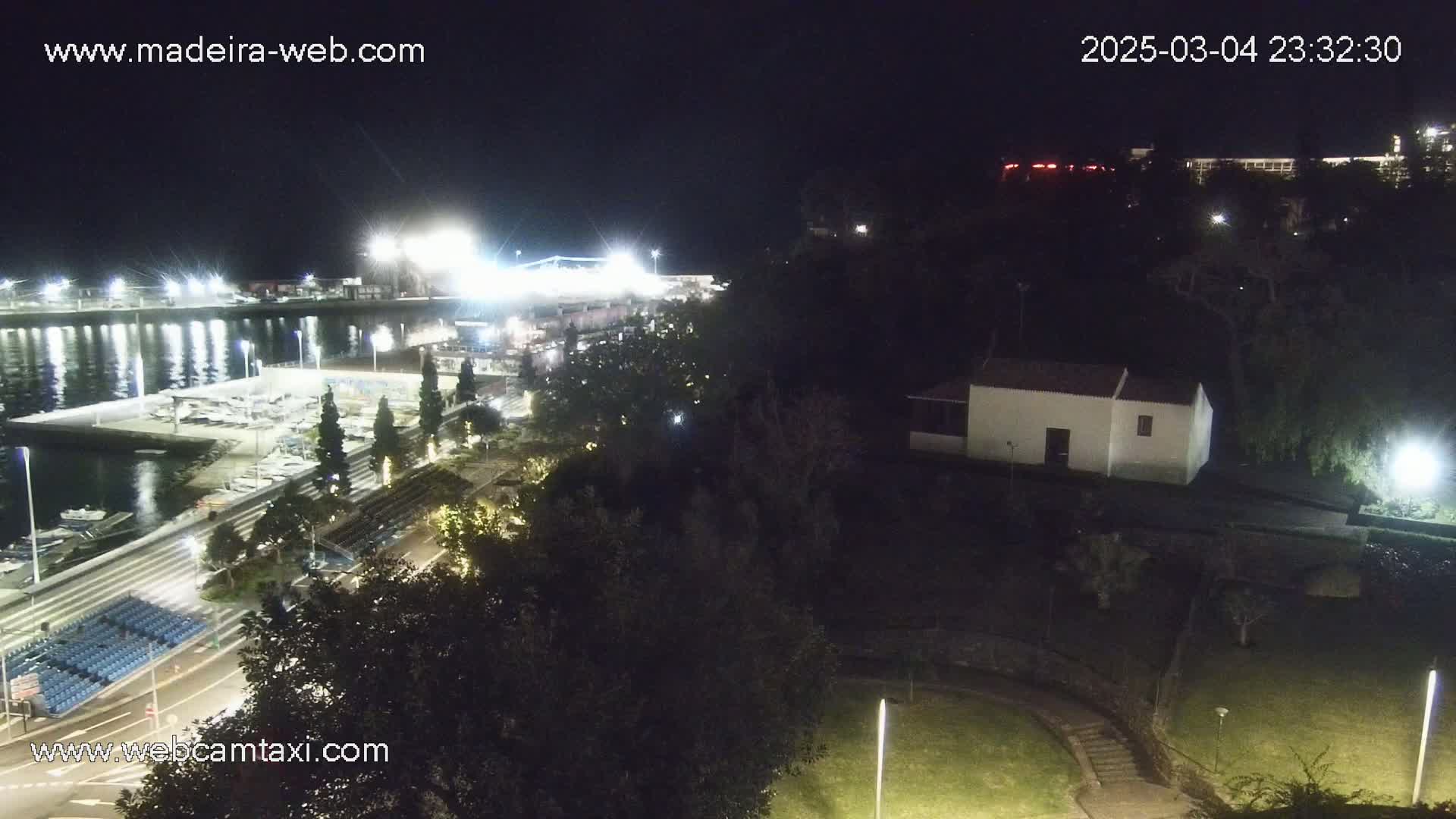 A nighttime, brightly lit marina with boats, a shoreline park, and a small white building situated on a hill overlooking the water.