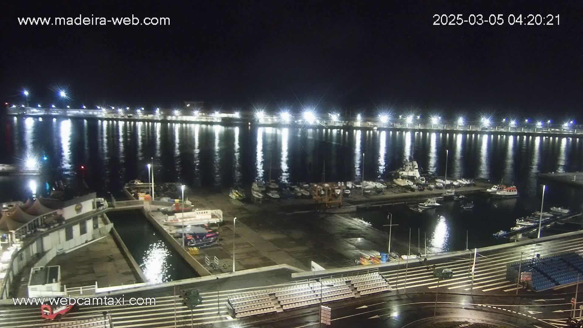A nighttime view of a harbor filled with boats, illuminated by bright lights reflecting on the wet pavement and calm water.