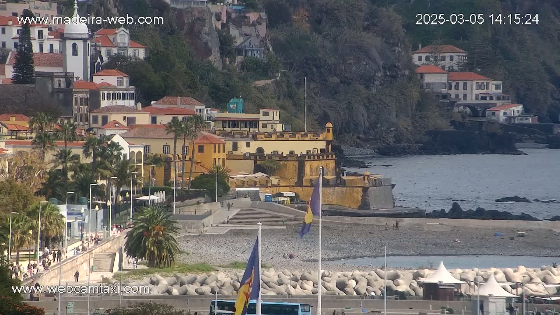 A coastal town with buildings on a hillside overlooking a pebble beach and calm ocean under a mostly clear sky.