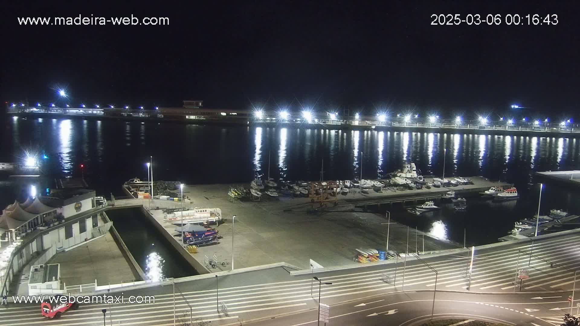 A nighttime view of a marina filled with docked boats, illuminated by bright lights reflecting on calm water.