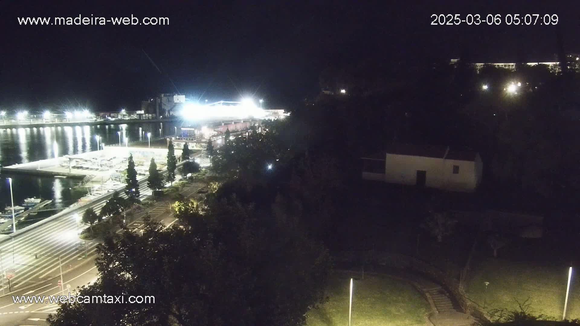 A nighttime view of a marina with boats docked, a road leading to brightly lit buildings, and dark trees and a small building on a hill overlooking the water.