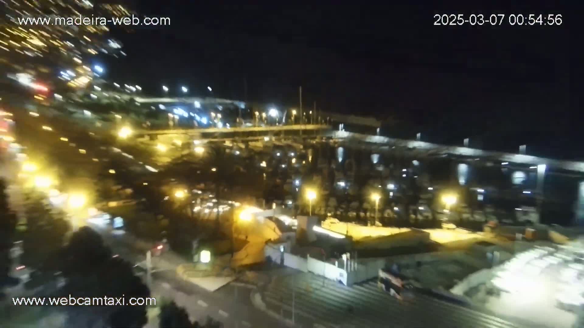 An aerial nighttime view shows a marina filled with boats, illuminated by streetlights and the glow of nearby buildings and traffic.