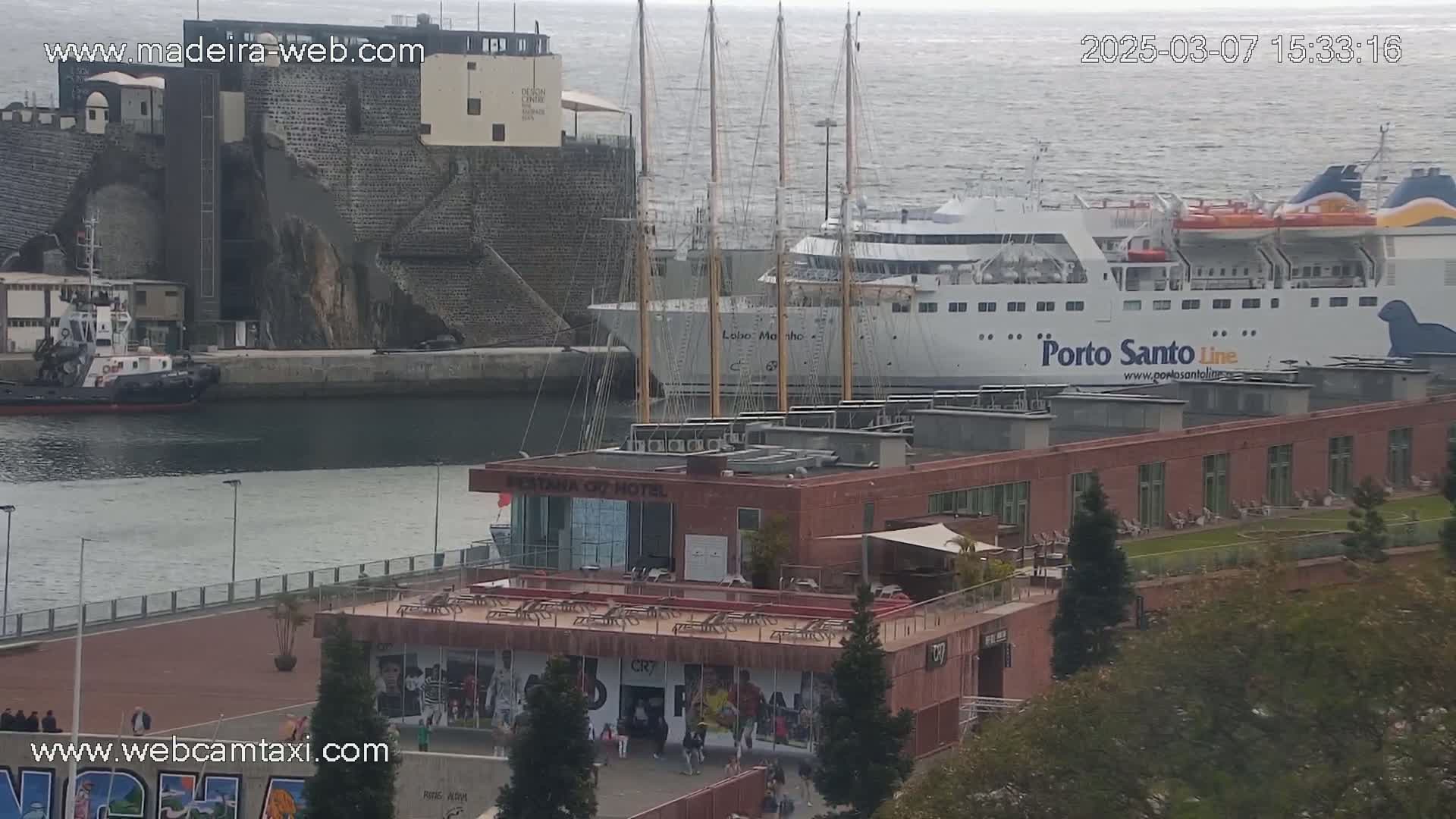 A harbor scene with a large ferry and a tall ship docked next to a brick building, on a partly cloudy day.