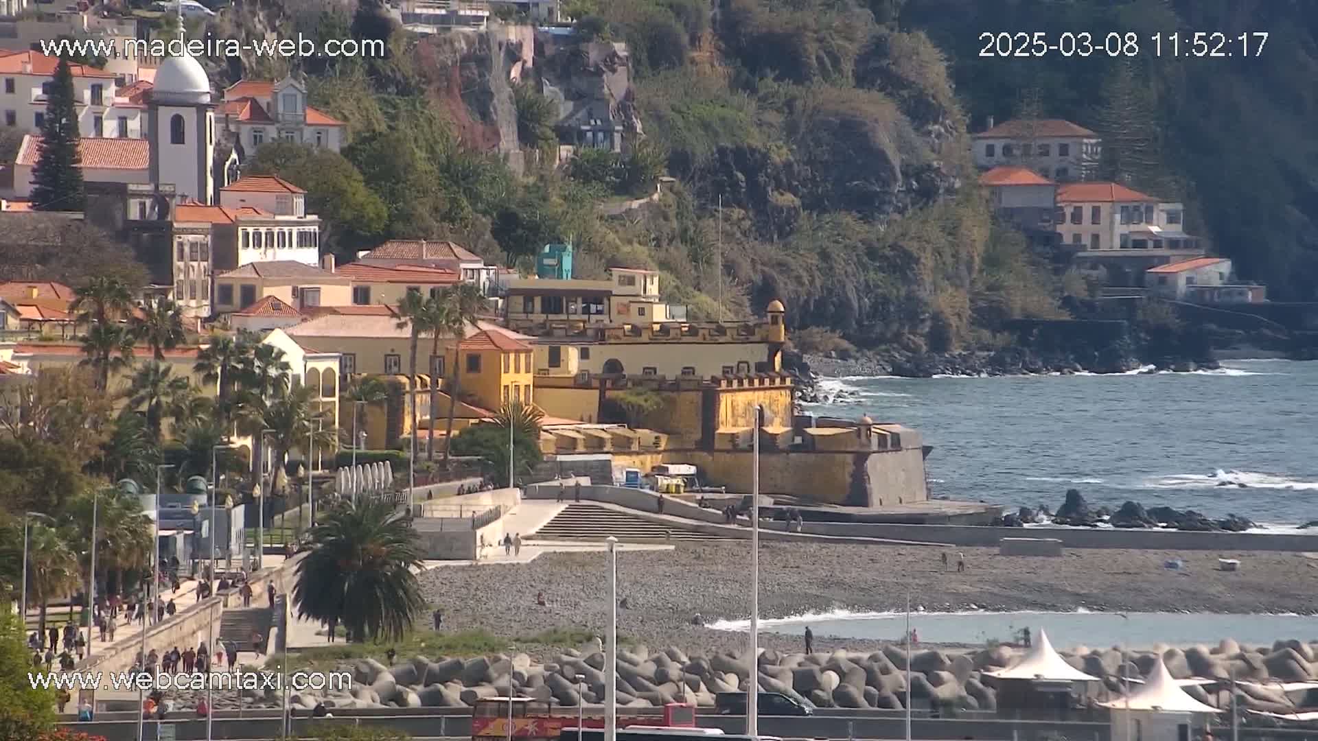 A seaside town with a yellowish fort, buildings with red tile roofs, palm trees, and a pebble beach on a sunny day with a calm ocean.