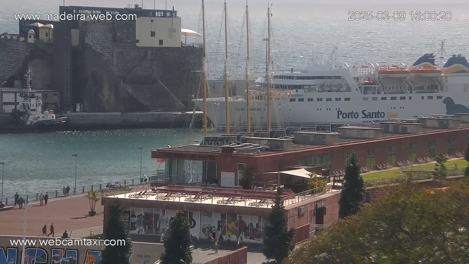 A harbor scene on a partly sunny day shows a large ferry, a tall sailing ship, a tugboat, a building with a rooftop patio, and a few people walking along a waterfront promenade.