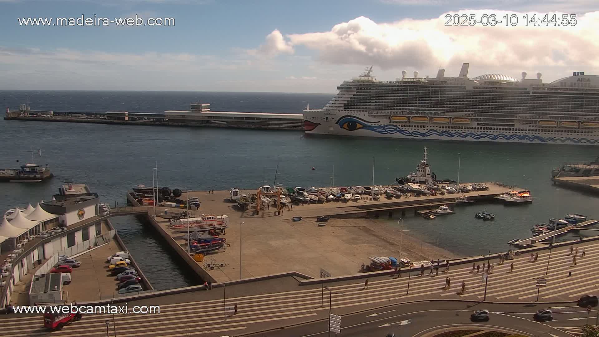 A large cruise ship is docked at a harbor on a partly cloudy day, with numerous smaller boats and people walking along a nearby promenade.