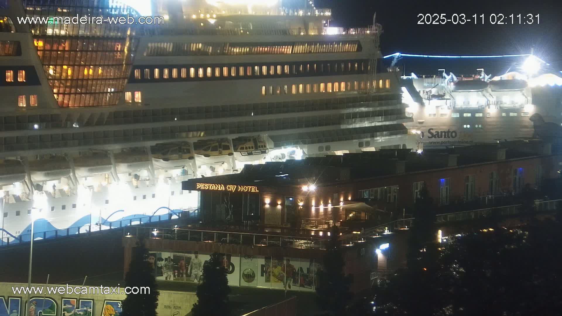 At night, a large cruise ship is docked next to a brightly lit hotel, with the surrounding area dimly lit.