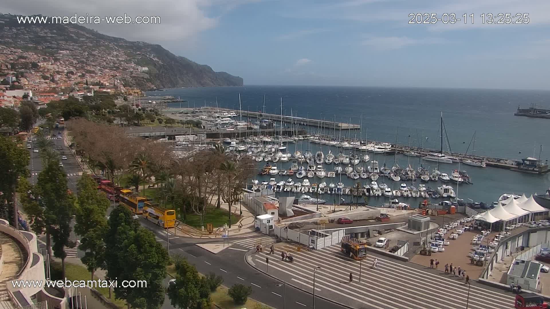 A harbor filled with numerous sailboats and yachts is situated next to a coastal town under a partly sunny sky.