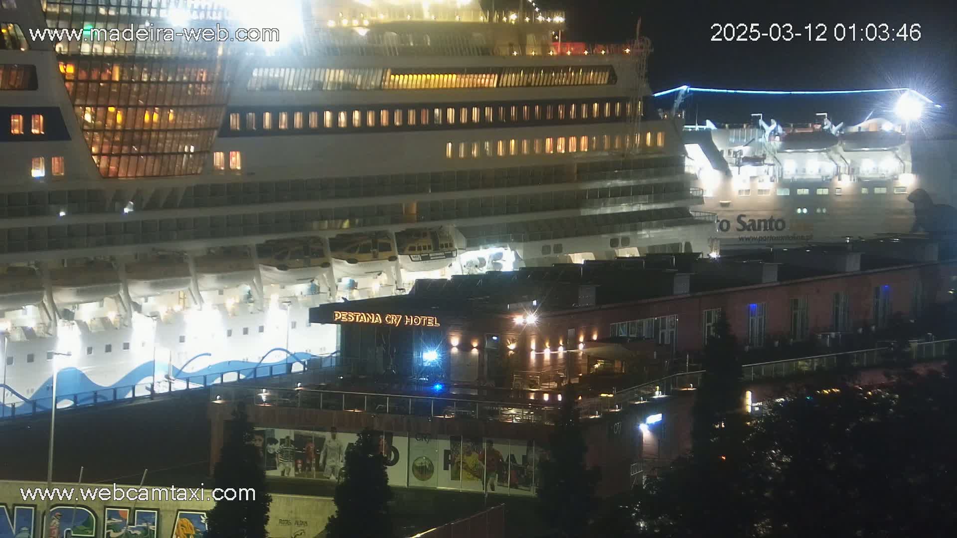 At night, a large cruise ship is docked next to a smaller ferry, near a hotel and other buildings.