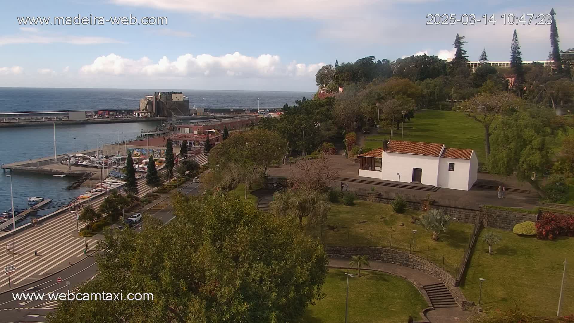 An aerial view of a harbor, marina, and park with a small white building, under partly cloudy skies.