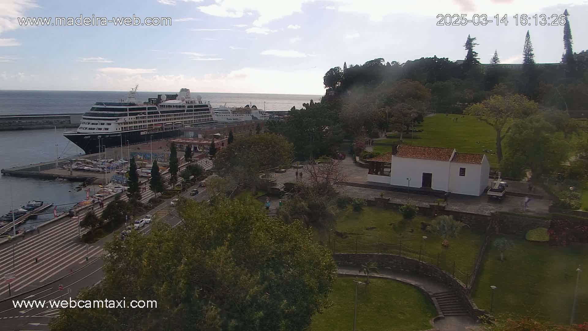 A large cruise ship is docked at a harbor, overlooking a park with a small white building and a grassy area, under a partly cloudy sky.