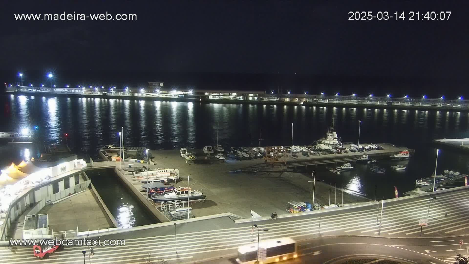 A nighttime view of a harbor filled with boats, a waterfront walkway, and a calm body of water reflecting lights from the shore.