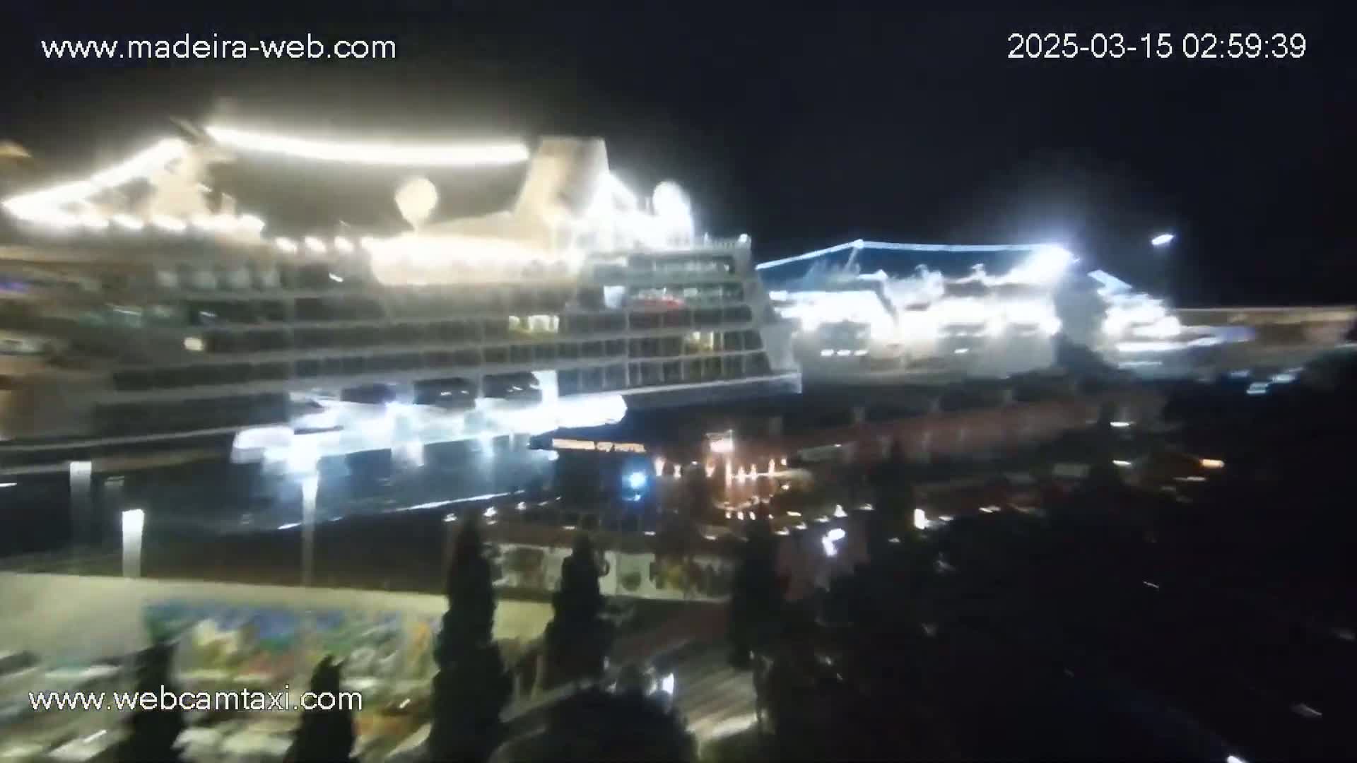 At night, two brightly lit cruise ships are docked, with a crowd of people gathered near the water's edge.