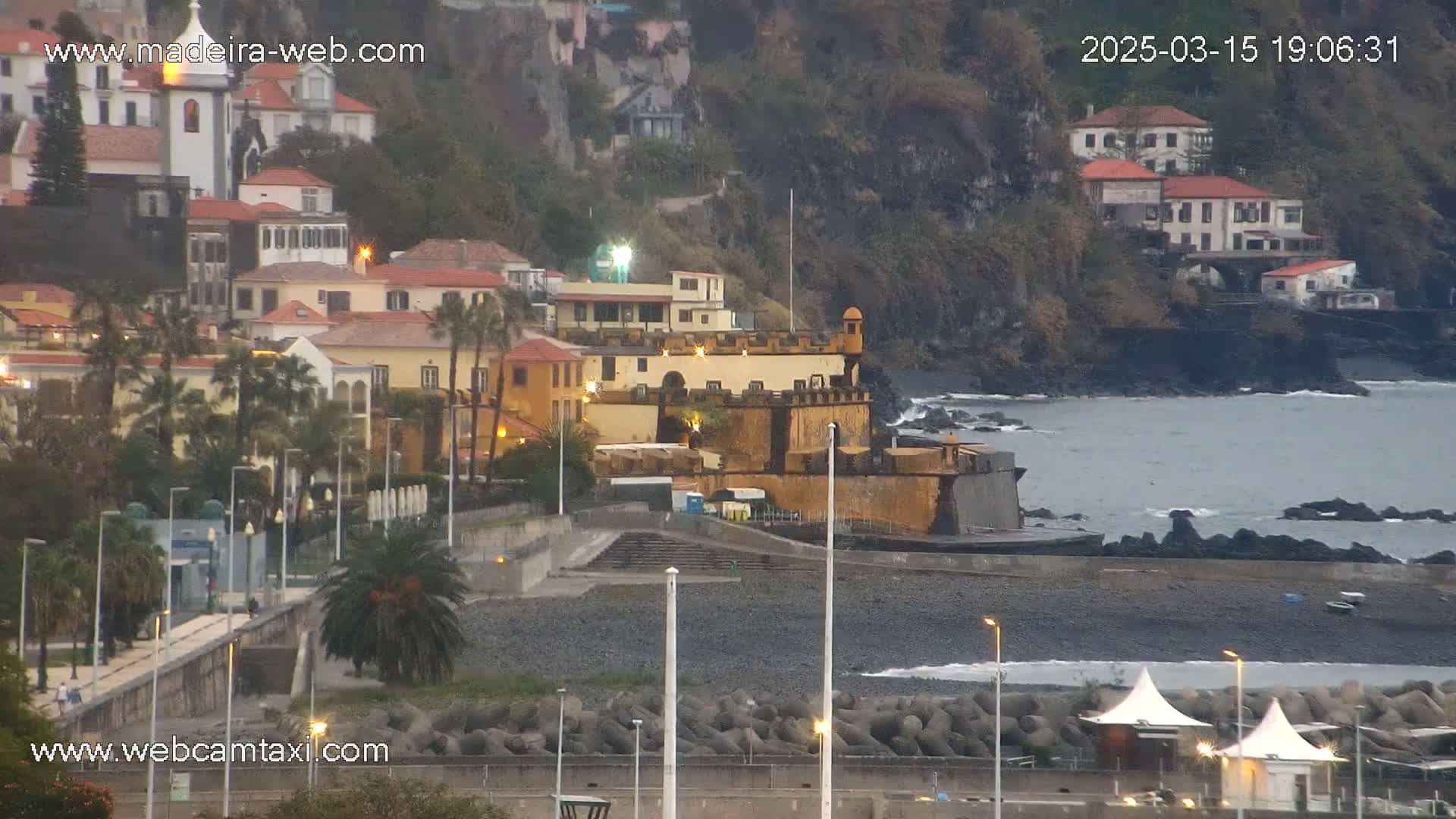 A coastal town with buildings on a hillside overlooking a dark-grey, rocky beach and calm ocean under an overcast sky.