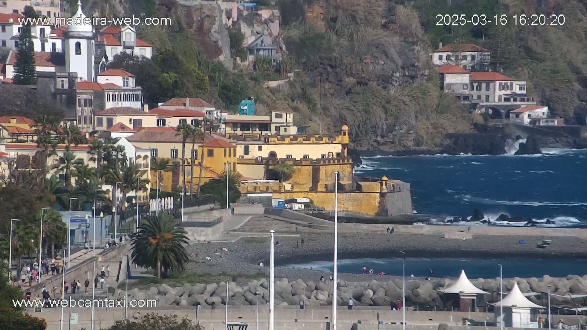 A coastal town with buildings on a hillside overlooking a rocky beach with moderate waves under sunny skies.