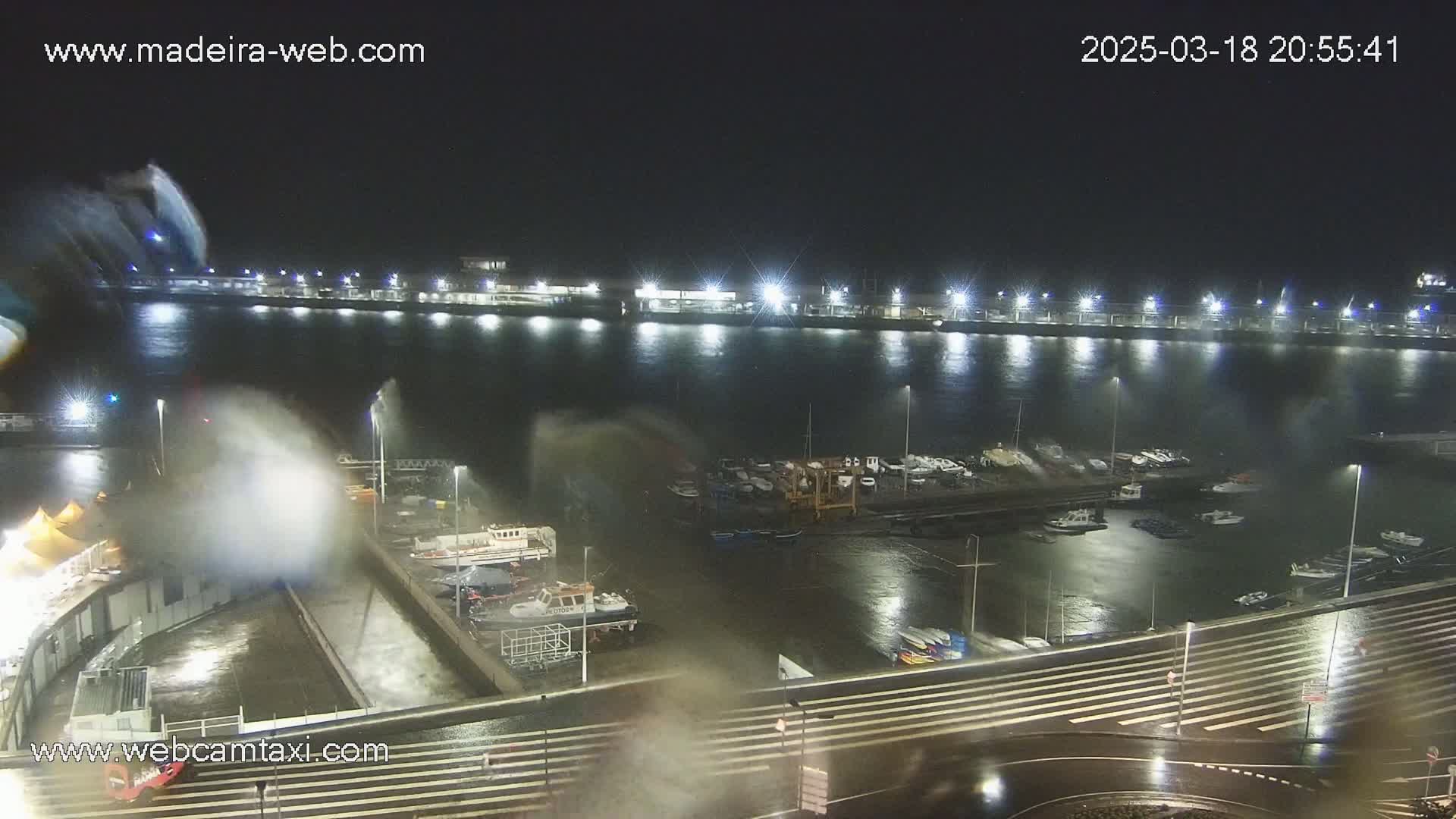 A nighttime view of a harbor with boats and a long pier, experiencing strong waves and spray under stormy conditions.