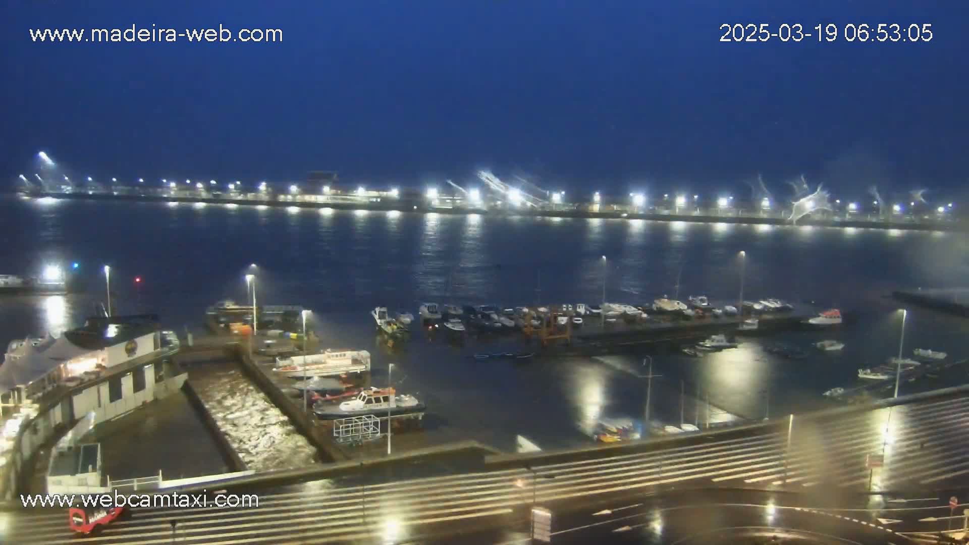 A nighttime harbor scene shows many boats moored in choppy water under a dark, possibly stormy sky.