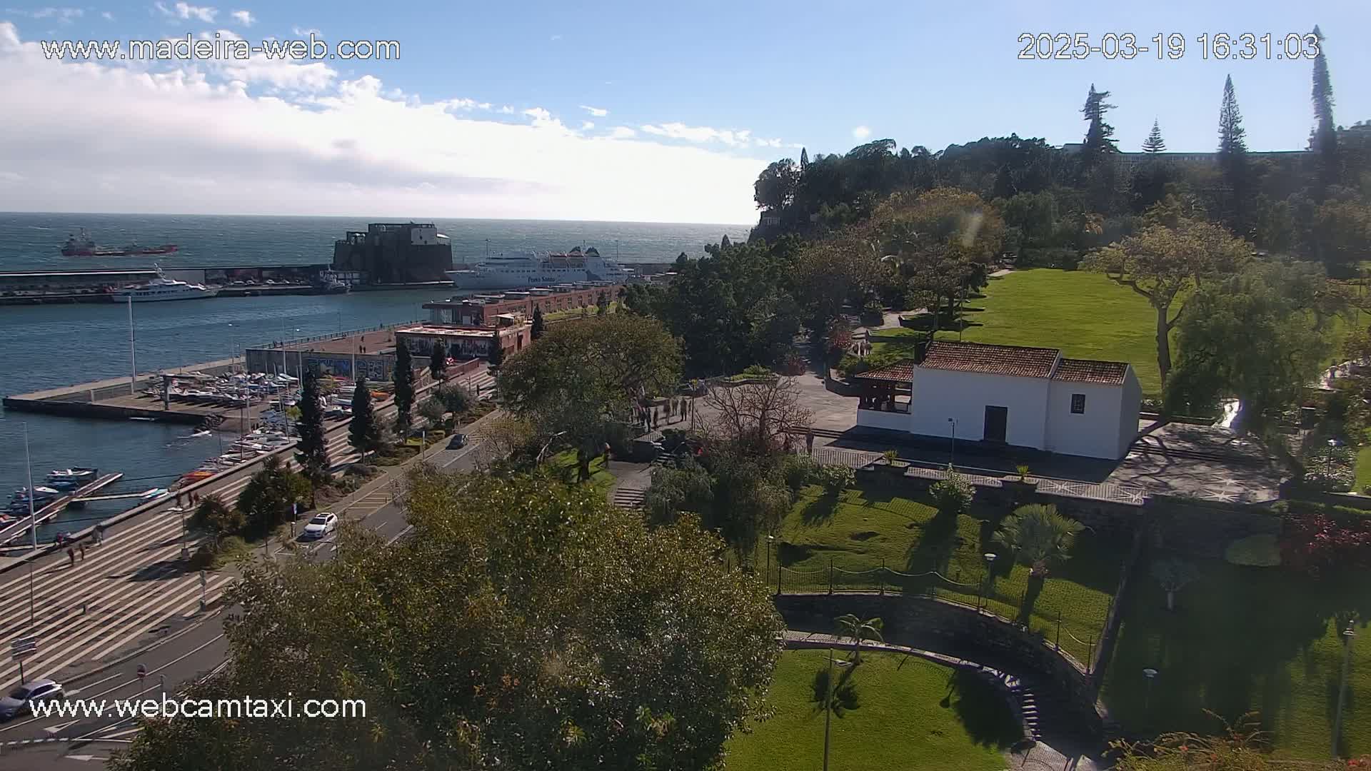 A harbor with boats and a ferry, next to a park with a small white building, under a mostly sunny sky.