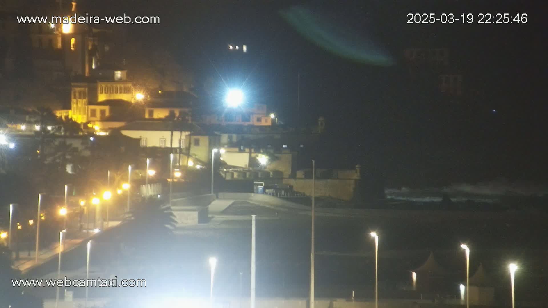 A nighttime view of a coastal town, illuminated by streetlights, with dark water and small waves visible in the distance.