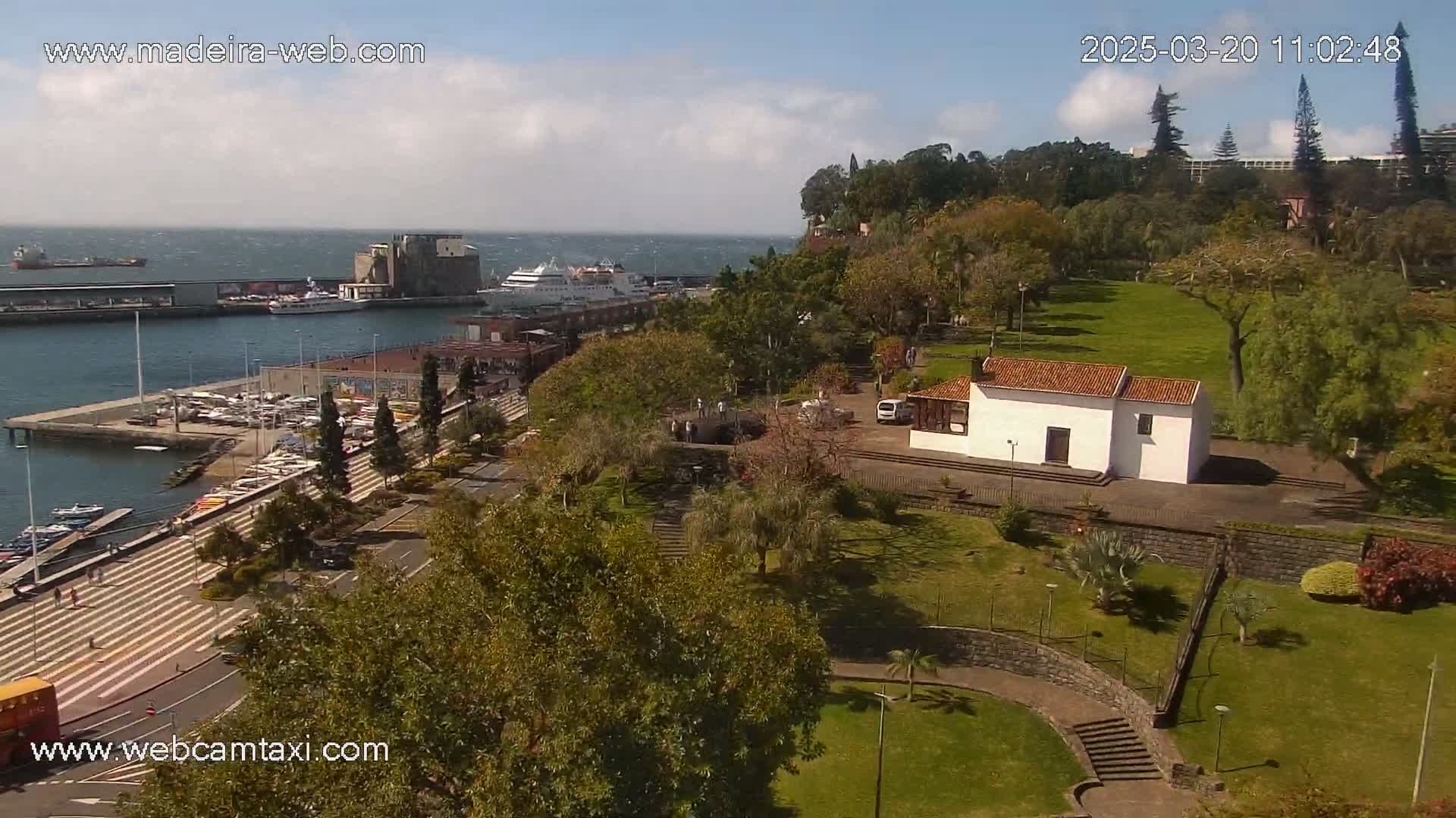 A harbor with boats and a cruise ship is visible in the distance, beside a park with a small white building and lush green lawns under a partly cloudy sky.