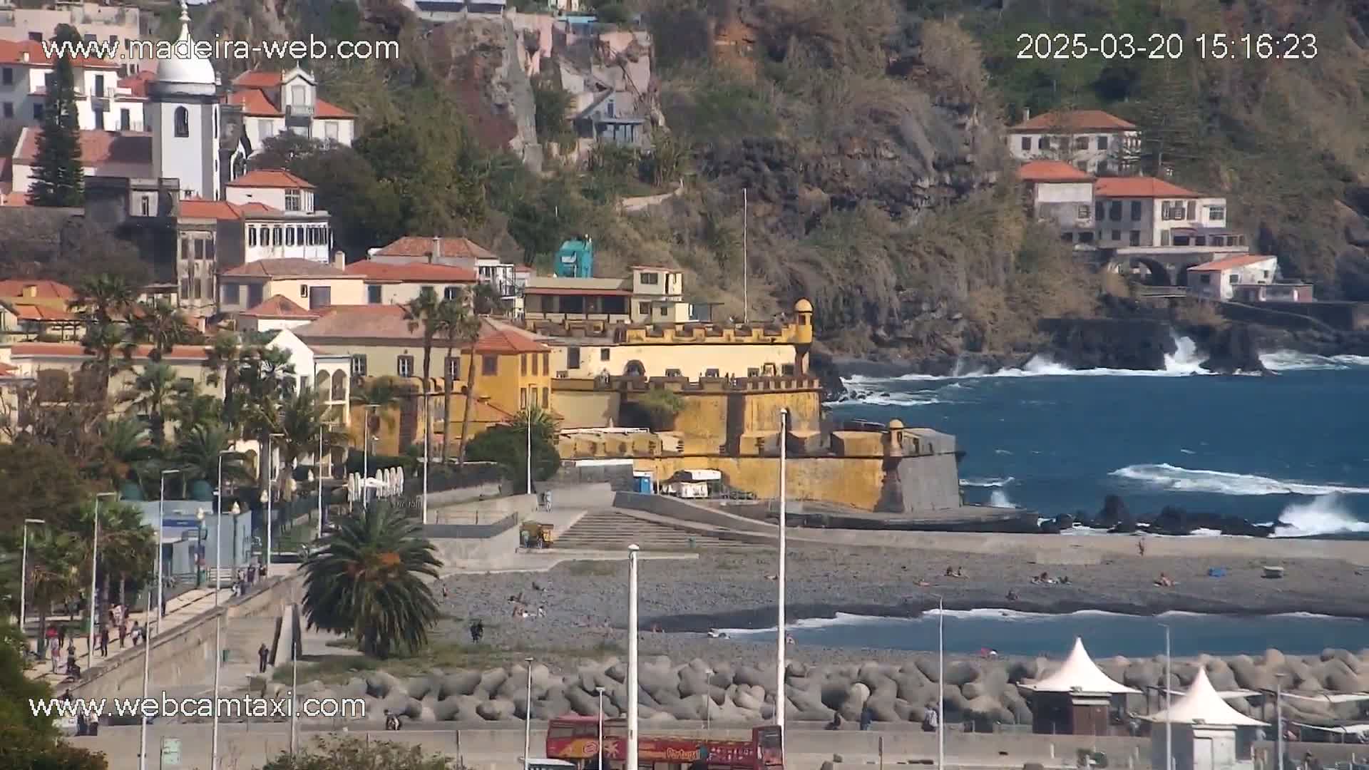 A coastal town with a yellow fortress sits on a rocky hillside overlooking a dark-sand beach and a blue ocean with moderate waves.