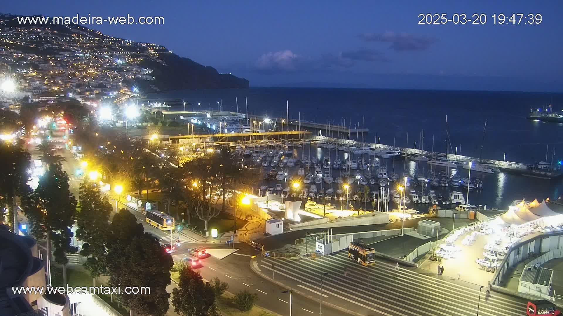 A nighttime view of a harbor filled with boats, adjacent to a city illuminated by streetlights, under a twilight sky with a few clouds.