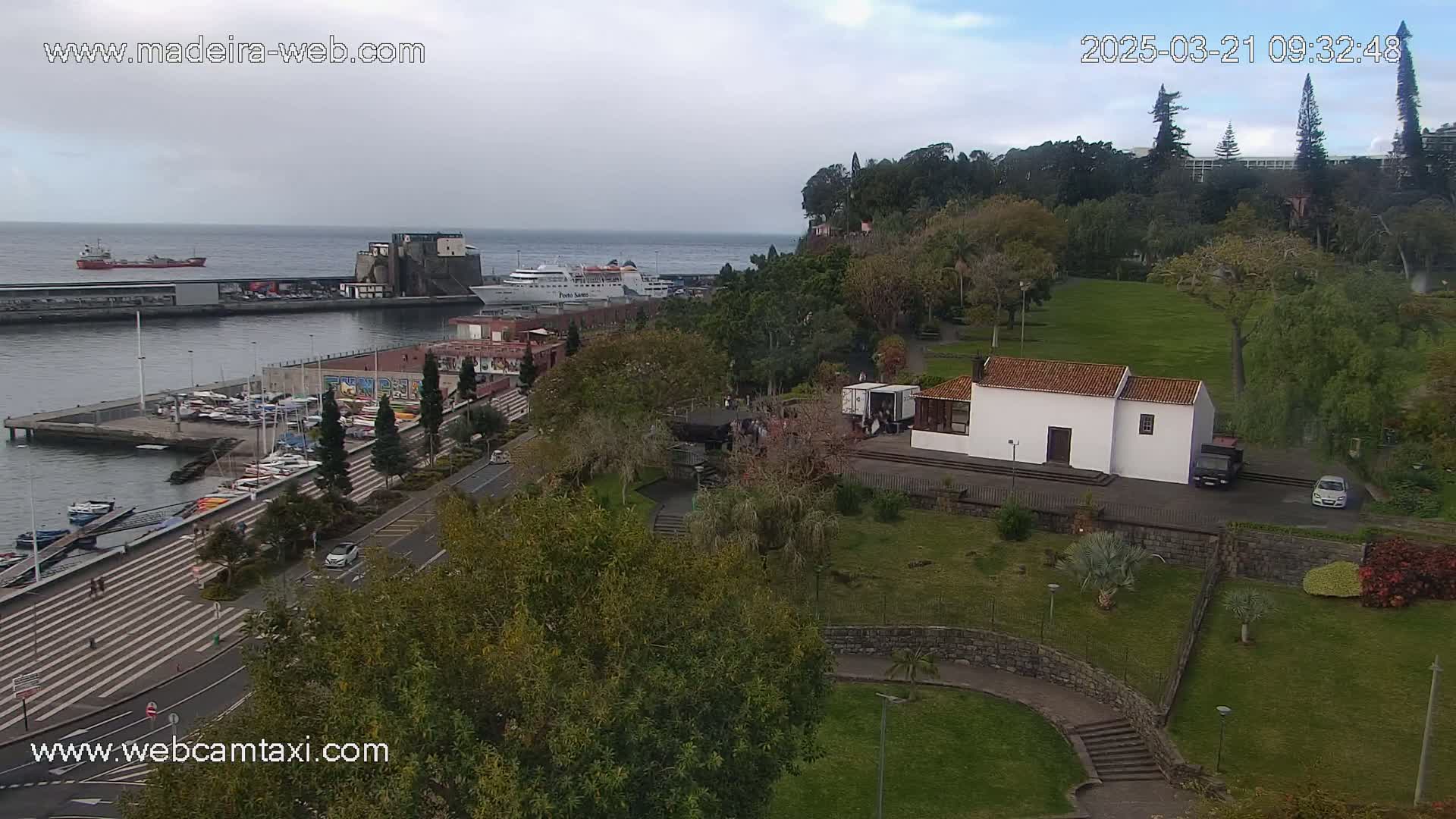 An elevated view of a harbor with boats, a waterfront promenade, and a park with a small white building under an overcast sky.
