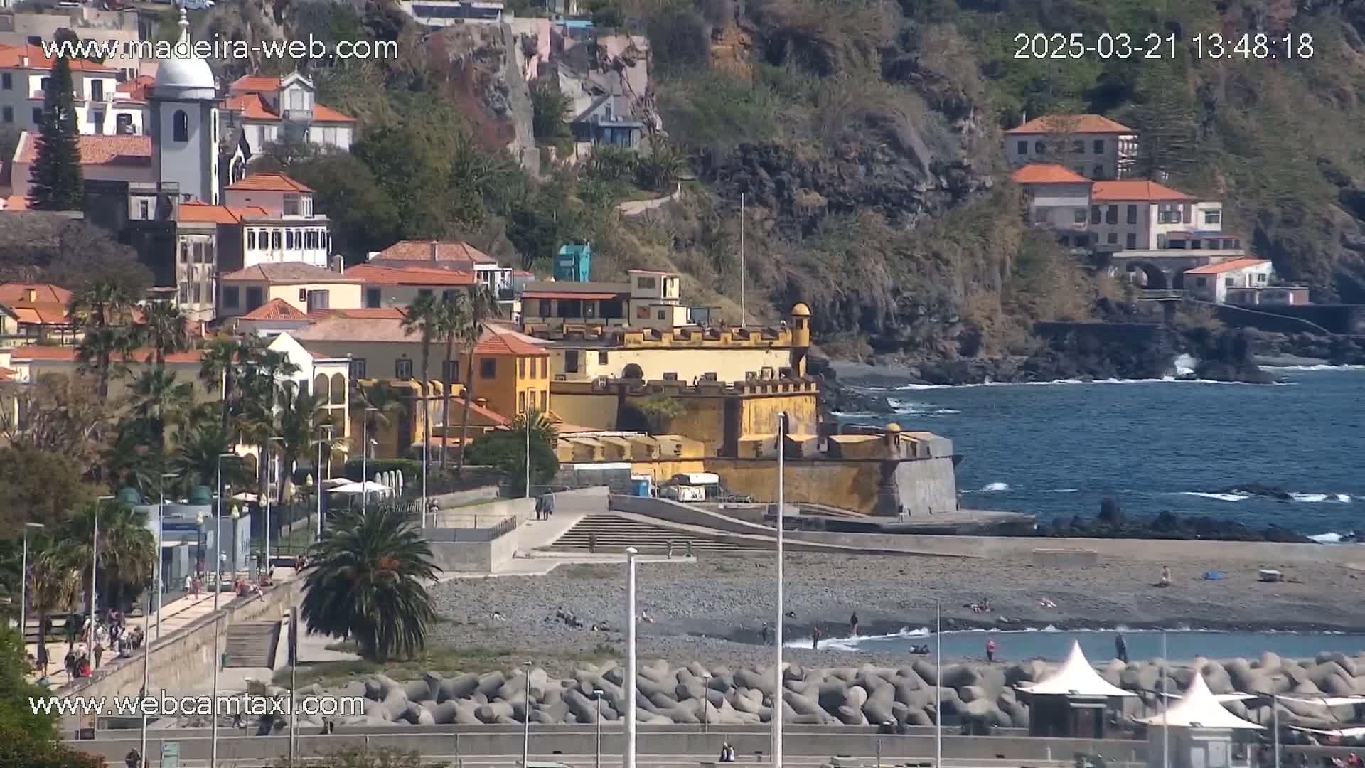 A coastal town with a yellow fort, buildings on a hillside, and a pebble beach with a few people, under sunny conditions.