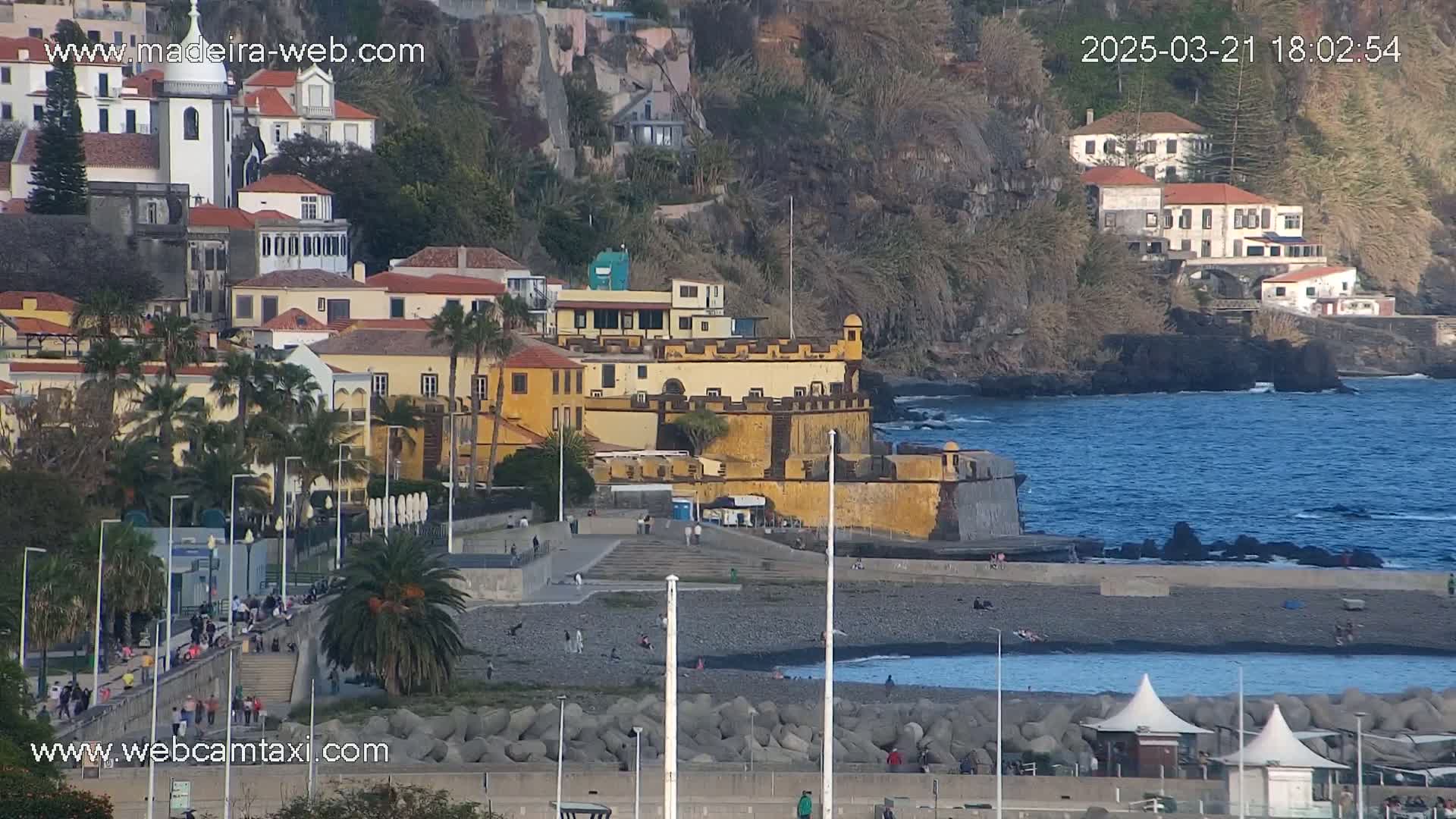 A coastal town with buildings and a fortress on a rocky shore, under a mostly clear sky.
