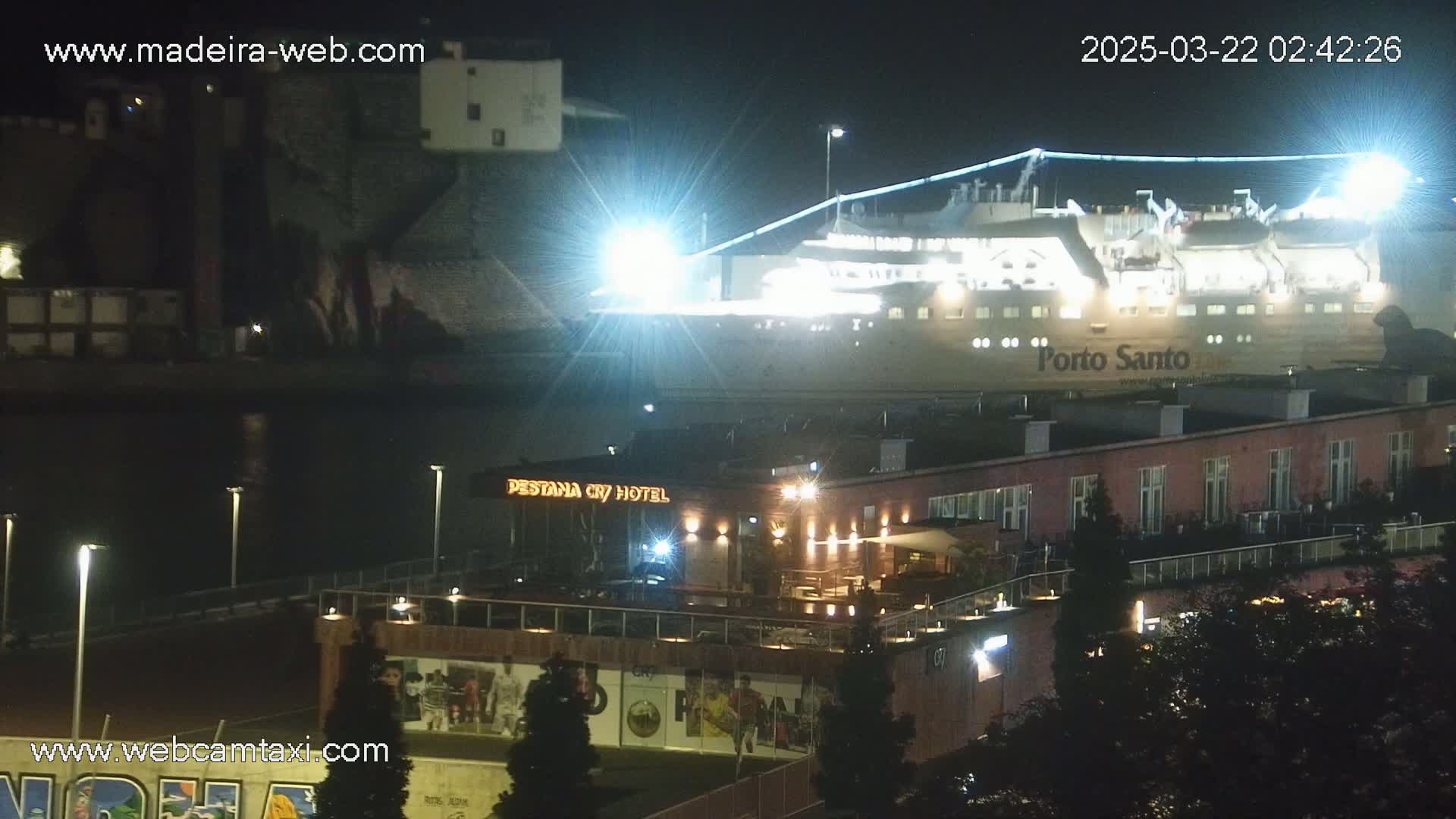 A large, brightly lit ferry is docked at night next to a hotel and other buildings.
